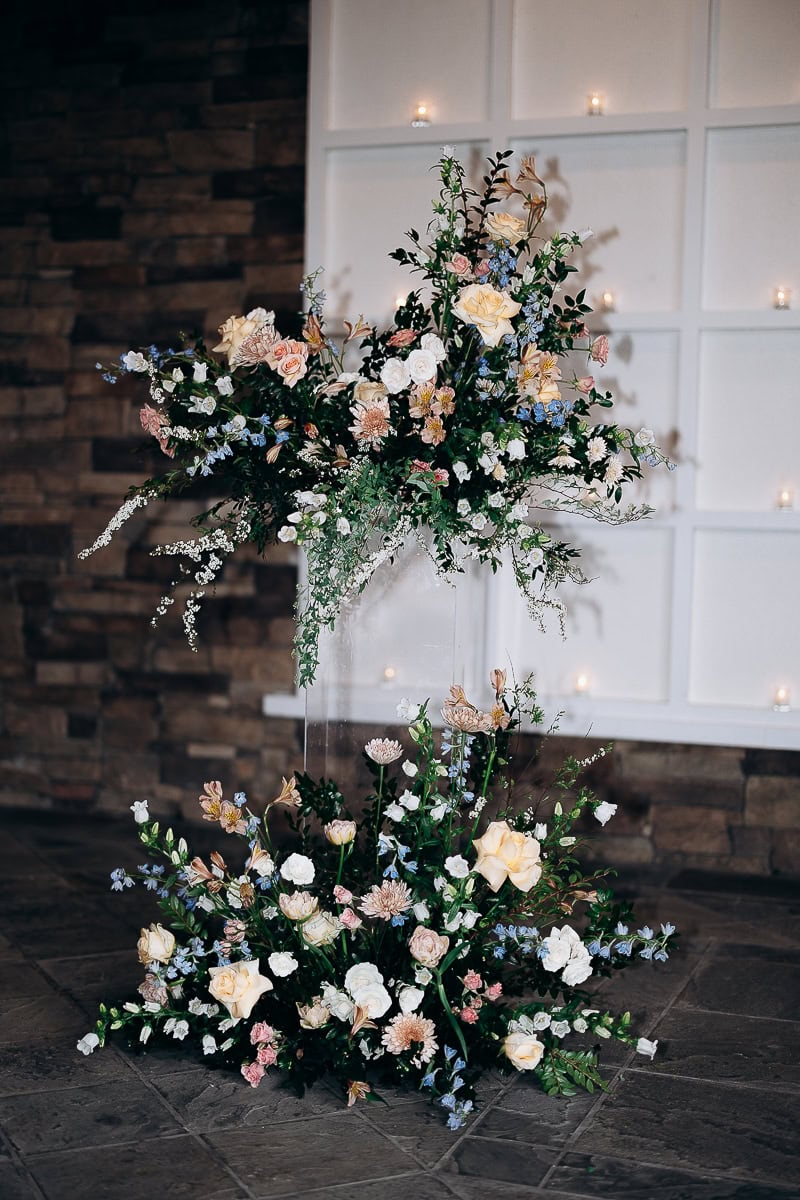 Tall and low floral arrangement with pastel roses, blue flowers, and greenery on stone floor and white paneled wall with candles at The Stone House, Warren NJ.