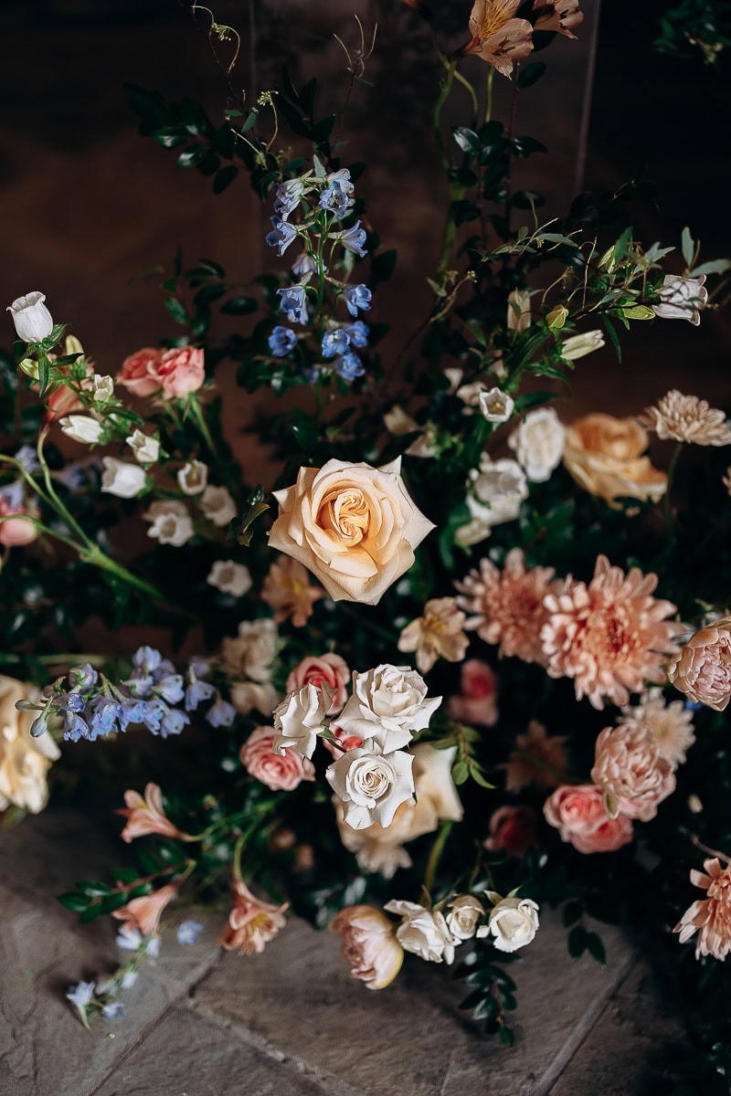 Close-up of a floral arrangement featuring cream and blush roses, blue delphiniums, and lush greenery on a stone floor.