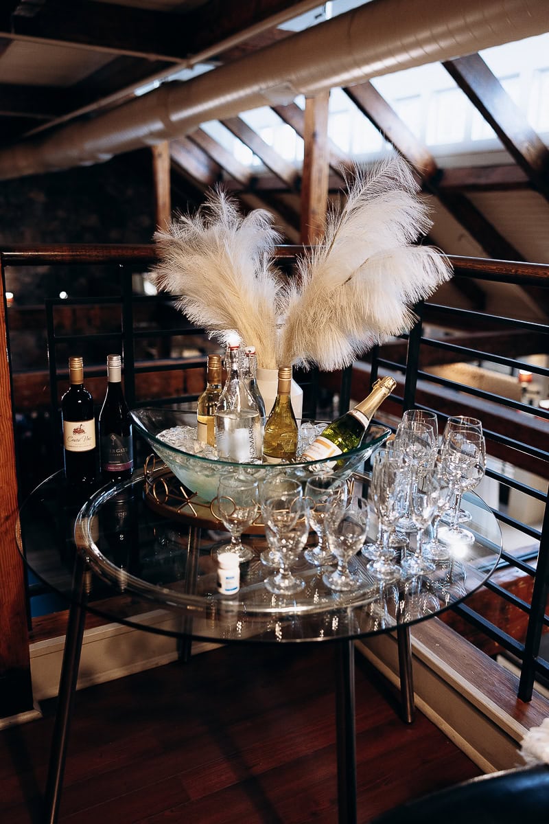 Glass table with ice-filled bowl holding bottles of wine and sparkling water, surrounded by empty wine glasses and pampas grass in a rustic loft setting.