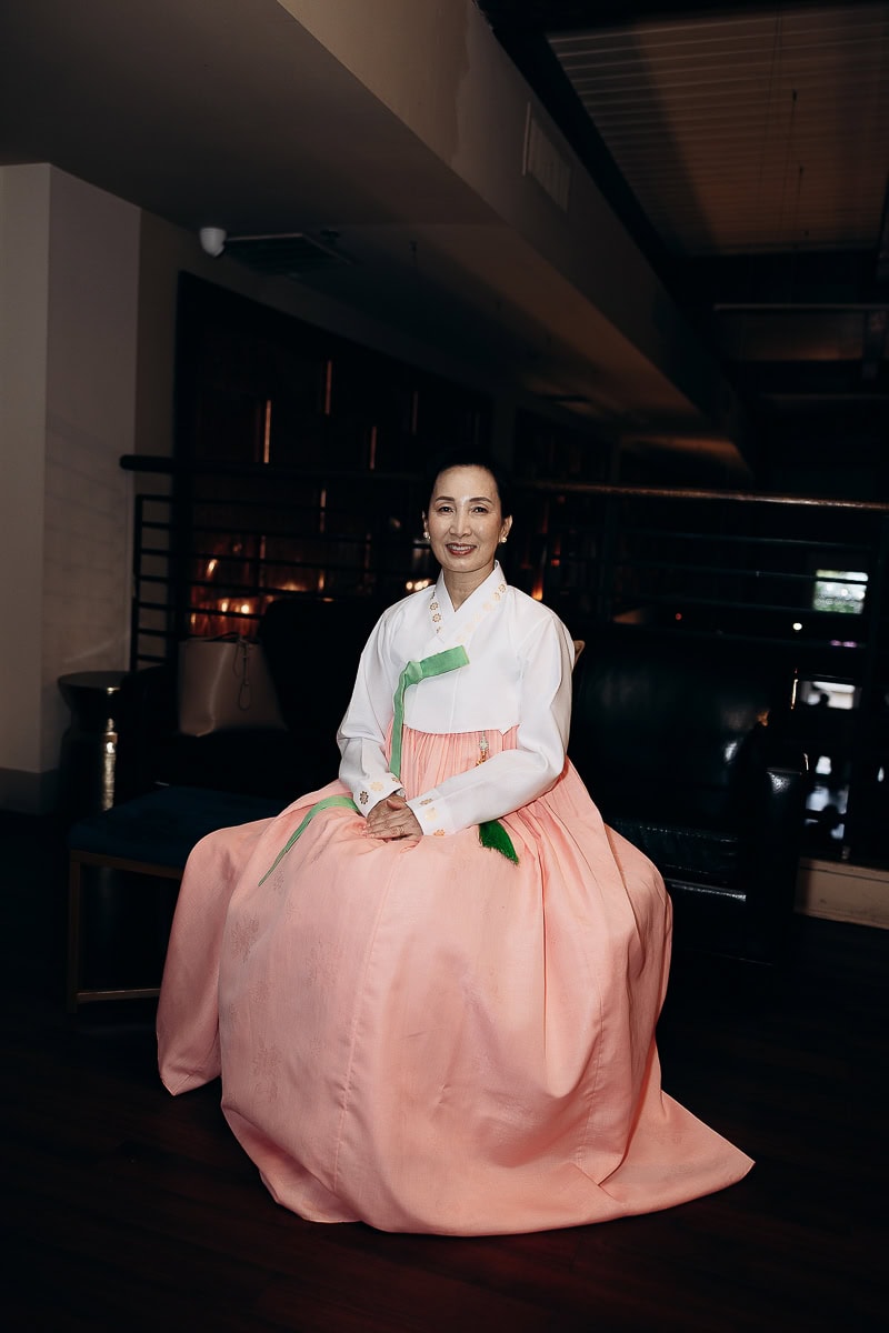 Woman seated indoors wearing a traditional peach and white hanbok with green accents, in a dimly lit room at The Stone House, Warren, NJ.