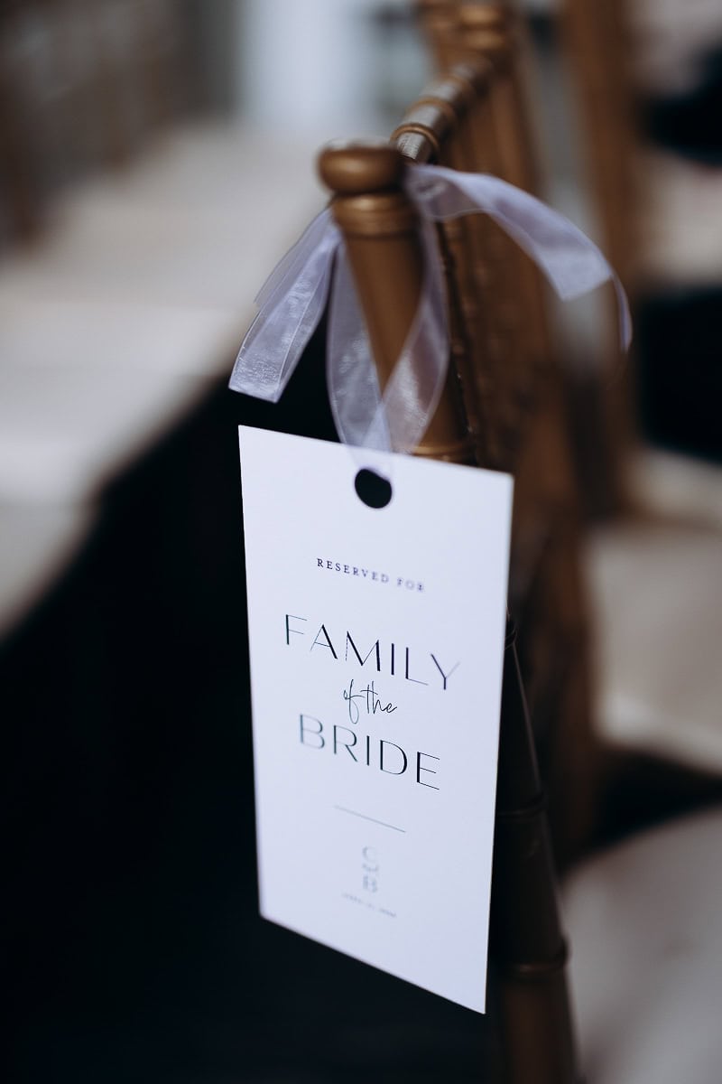 Close-up of a white reserved sign reading 'Family of the Bride' tied with a sheer ribbon to a gold Chiavari chair at a wedding venue.