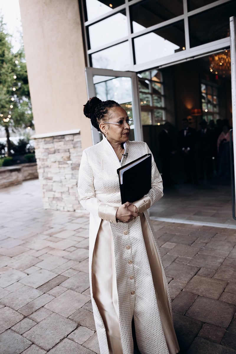 Woman in ivory textured long coat holding a black portfolio standing outside a venue with glass doors, The Stone House, Warren, NJ.