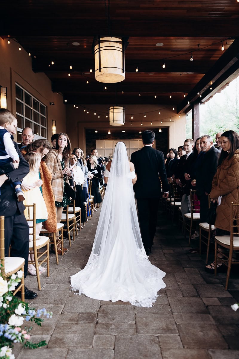 Bride in flowing white gown and veil walking arm-in-arm with groom in black suit down aisle lined with seated guests under warm lanterns and string lights at The Stone House, Warren, NJ.