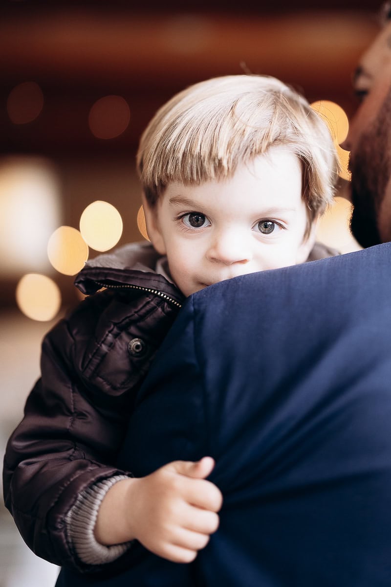 Close-up of a young child with light hair holding onto an adult's shoulder, with warm bokeh lights in the background at The Stone House, Warren, NJ.