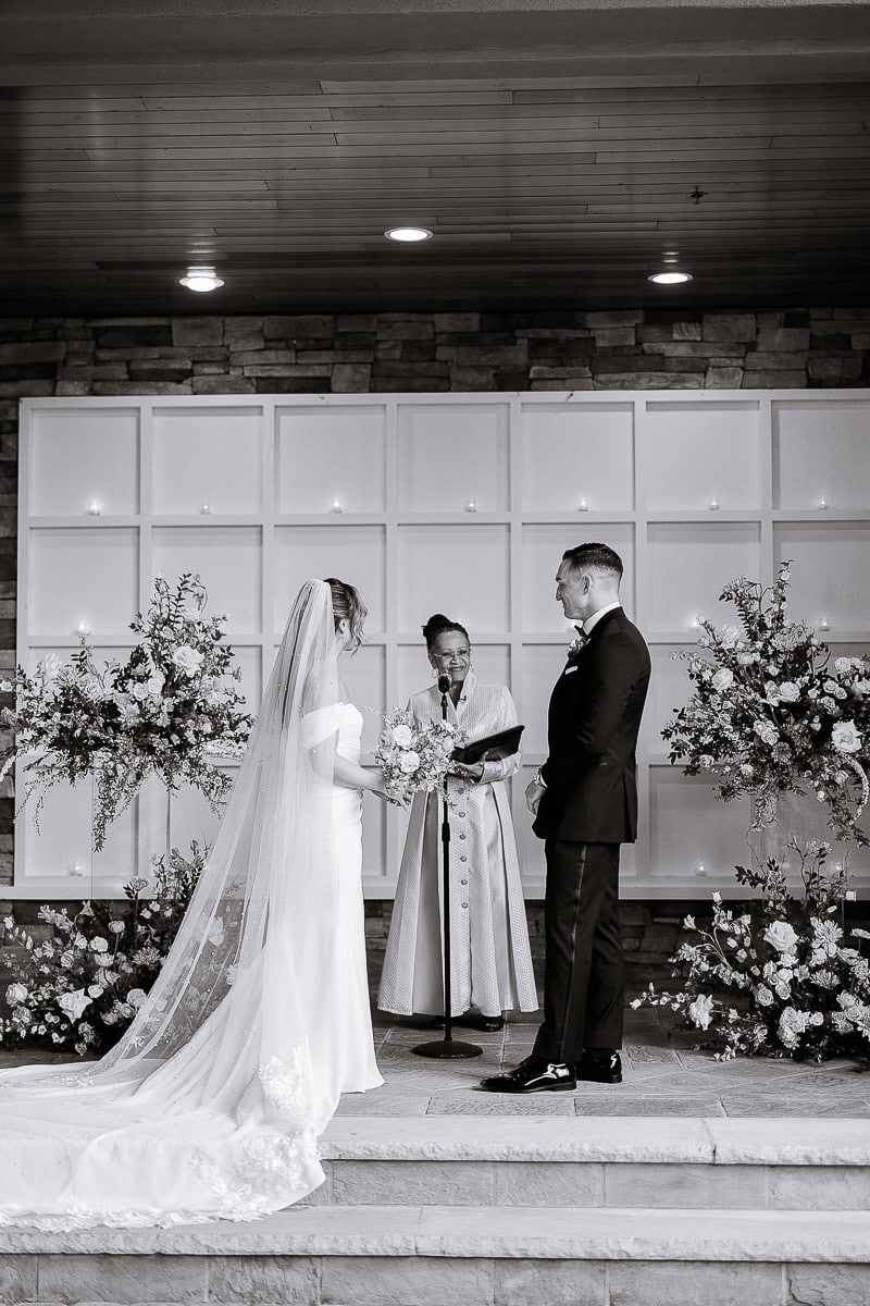 Bride and groom standing before officiant during wedding ceremony at The Stone House, Warren NJ, surrounded by floral arrangements and candles.