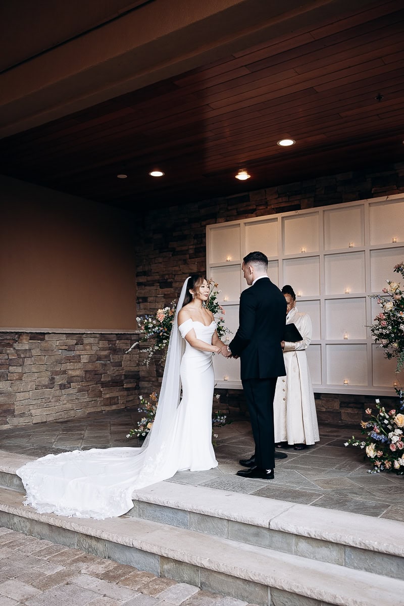 Bride in off-shoulder white gown with long train and veil holding hands with groom in black suit during wedding ceremony at The Stone House, Warren NJ