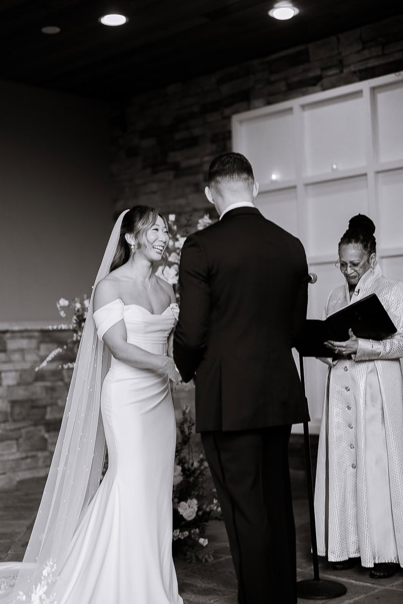 Bride in off-shoulder wedding gown smiling at groom during vow exchange with officiant reading from book, indoors at The Stone House, Warren NJ.