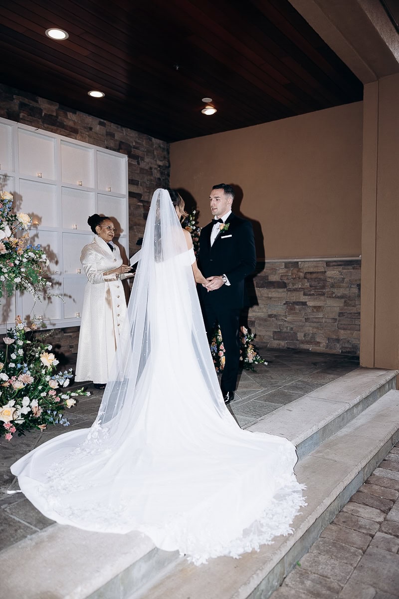 Bride in flowing white gown and veil holding hands with groom in black tuxedo during wedding ceremony at The Stone House, Warren NJ, with officiant and floral decor.