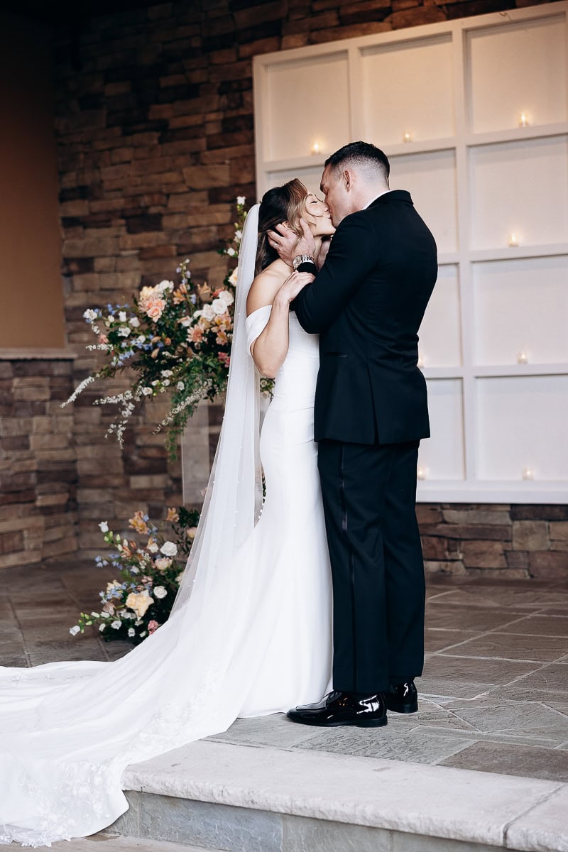 Bride and groom sharing a kiss indoors against a stone wall with floral arrangements and soft candlelight at The Stone House, Warren, NJ.