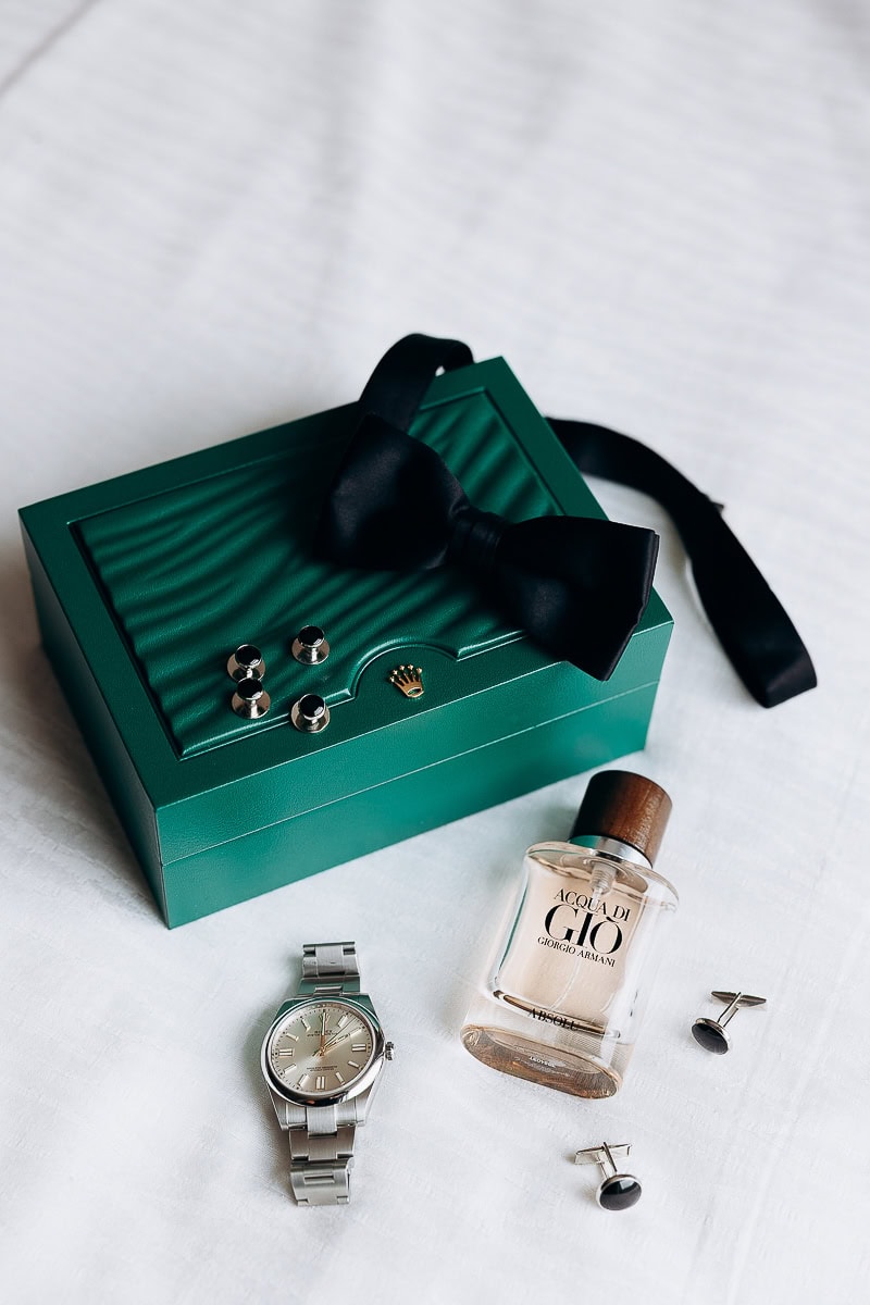Black silk bow tie, silver cufflinks, stainless steel Rolex watch, and Giorgio Armani Acqua di Giò perfume bottle on green Rolex box on white fabric.