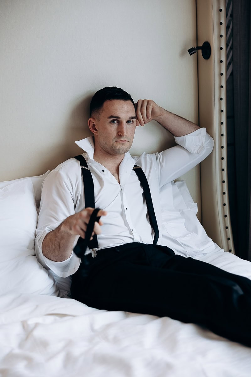 Man in white dress shirt and black suspenders reclining on a white bed against a cream headboard, looking thoughtfully at the camera.