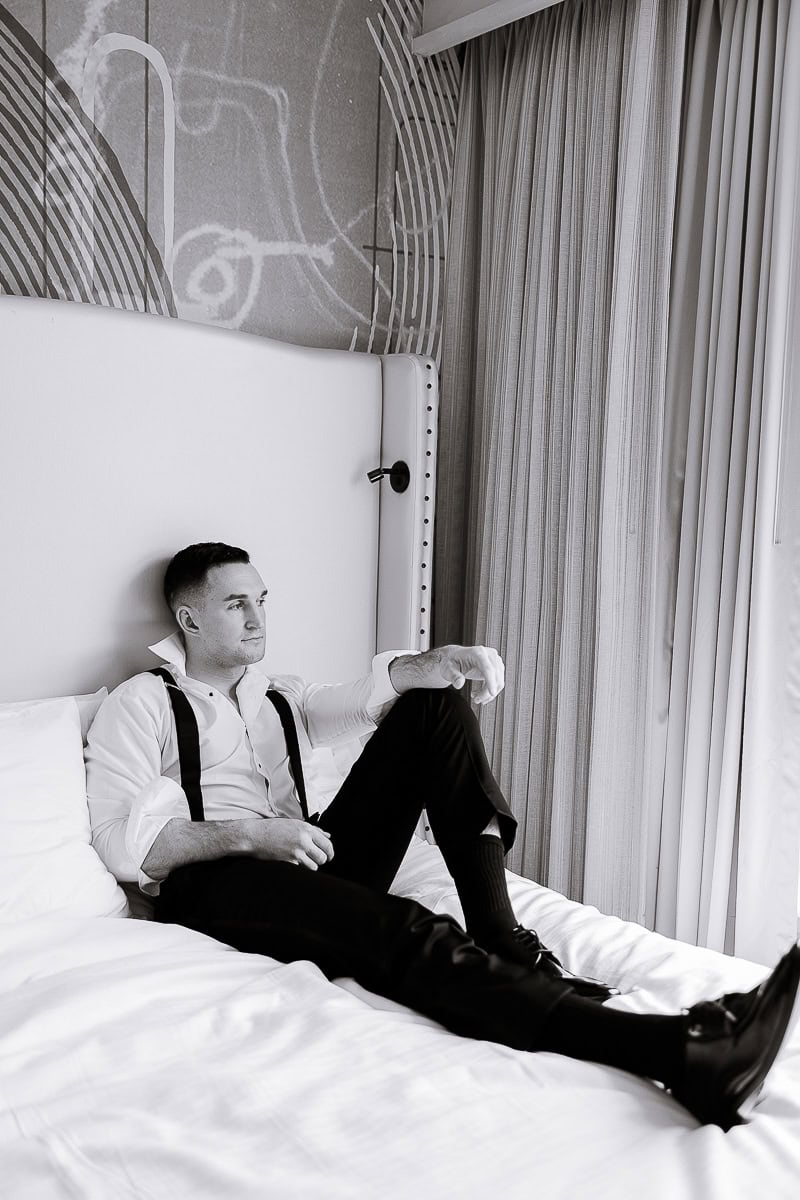Man in white dress shirt with suspenders sitting on bed, looking contemplative, with abstract wall art and curtains in background at The Stone House, Warren, NJ.