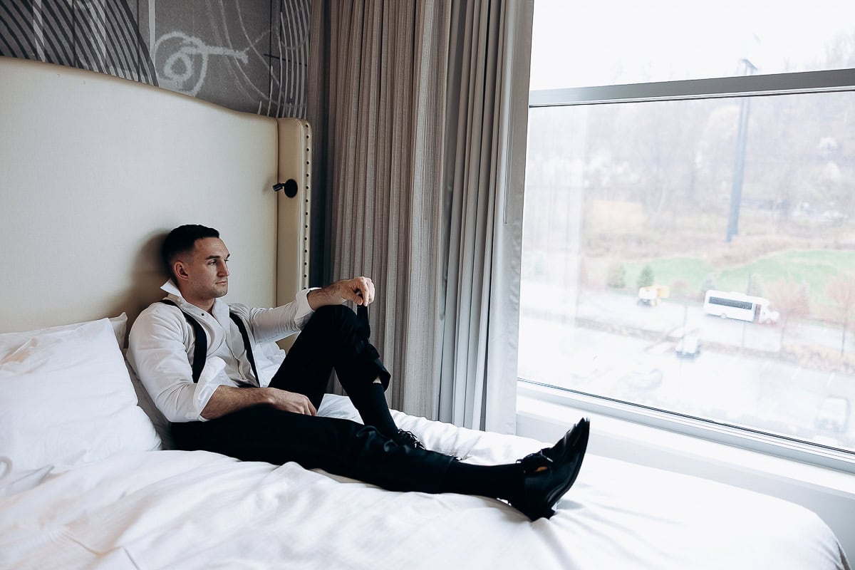Man in white shirt and black suspenders sitting on a white bed, looking pensively out a large window in a modern hotel room at The Stone House, Warren, NJ.