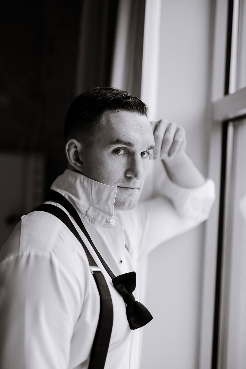 Black and white portrait of a groom wearing a white dress shirt with suspenders and an undone black bow tie, leaning against a window at The Stone House, Warren, NJ.