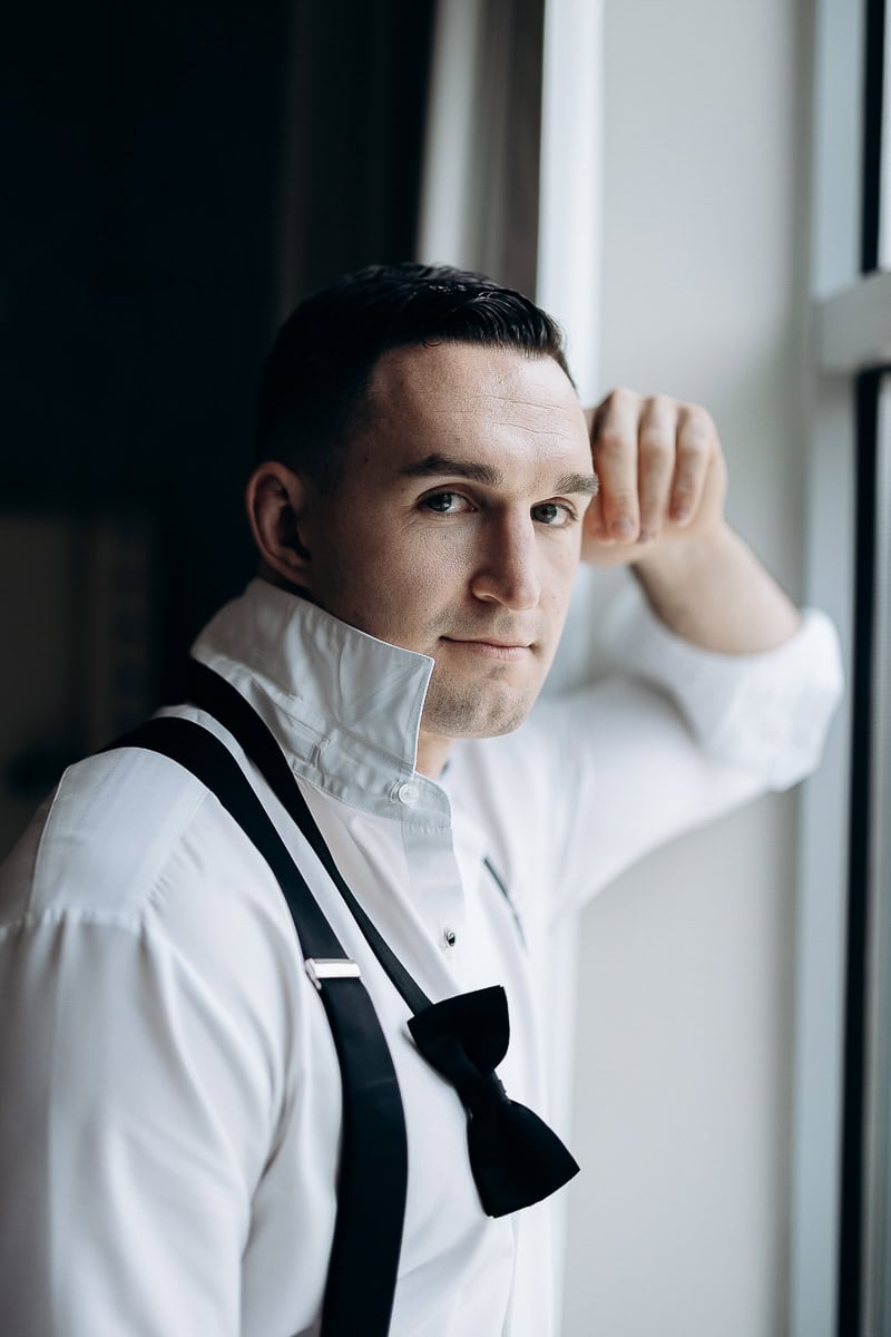 Man in white dress shirt with black suspenders and untied black bow tie, leaning against a window with soft natural light