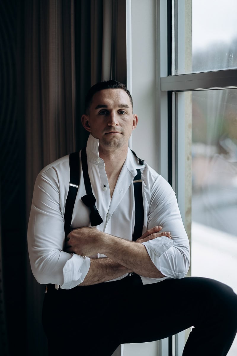 Man in white dress shirt with black suspenders sitting by a window, arms crossed, looking directly at camera in soft natural light.