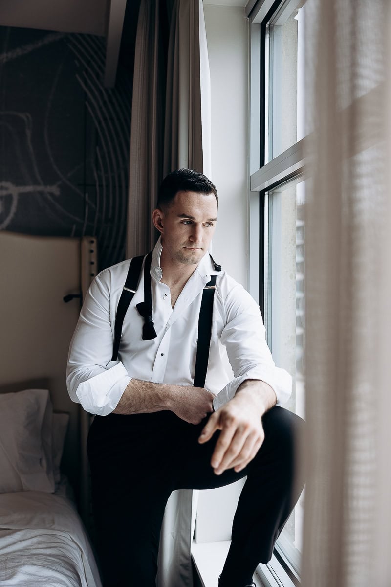 Man in white dress shirt with black suspenders sitting by a window, looking contemplative in a softly lit room.