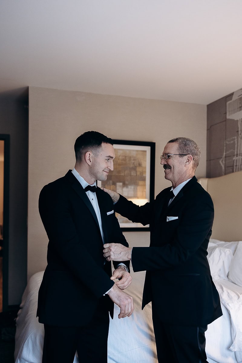 Groom and older man in tuxedos sharing a moment while adjusting suit in a softly lit hotel room at The Stone House, Warren, NJ.