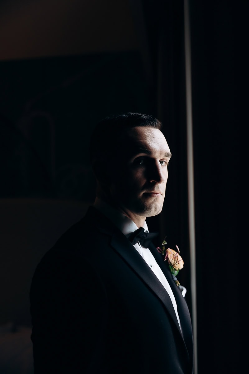Man in black tuxedo with bow tie, illuminated by window light, standing indoors in low light at The Stone House, Warren, NJ.