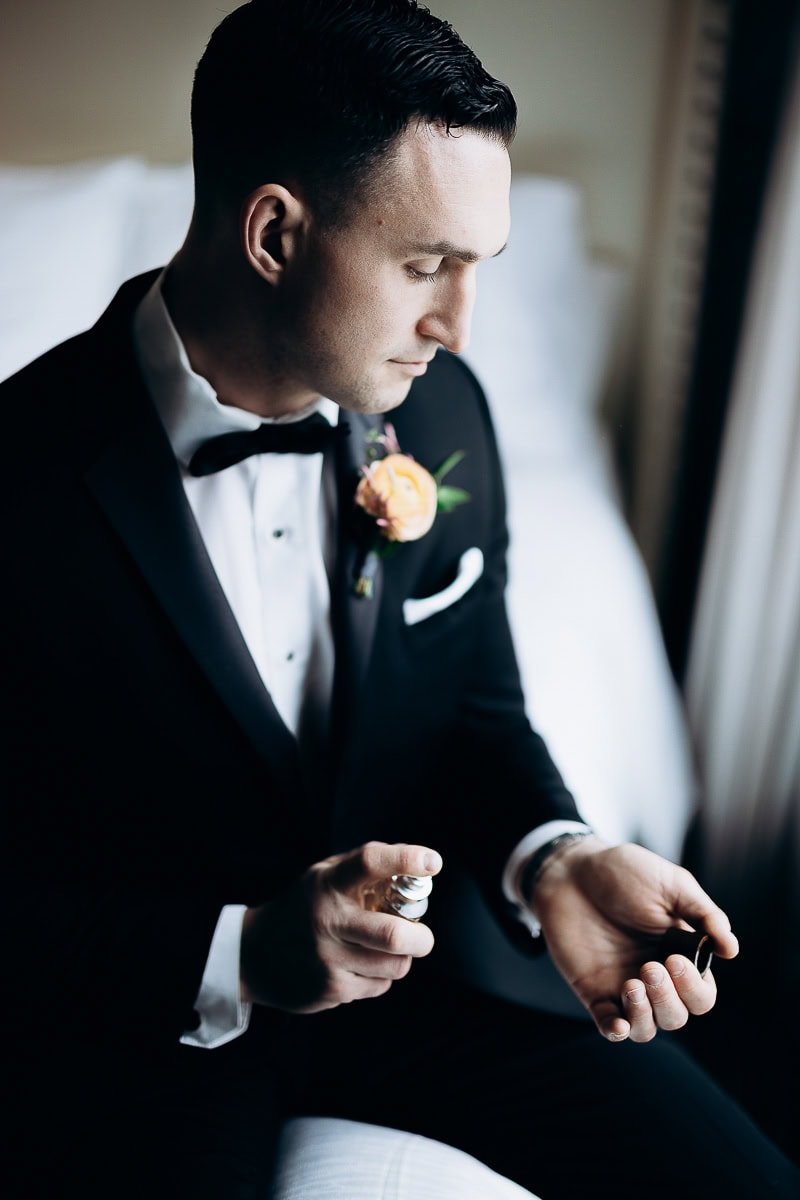 Groom in black tuxedo sitting on bed applying cologne, with peach boutonniere, soft natural light at The Stone House, Warren, NJ.