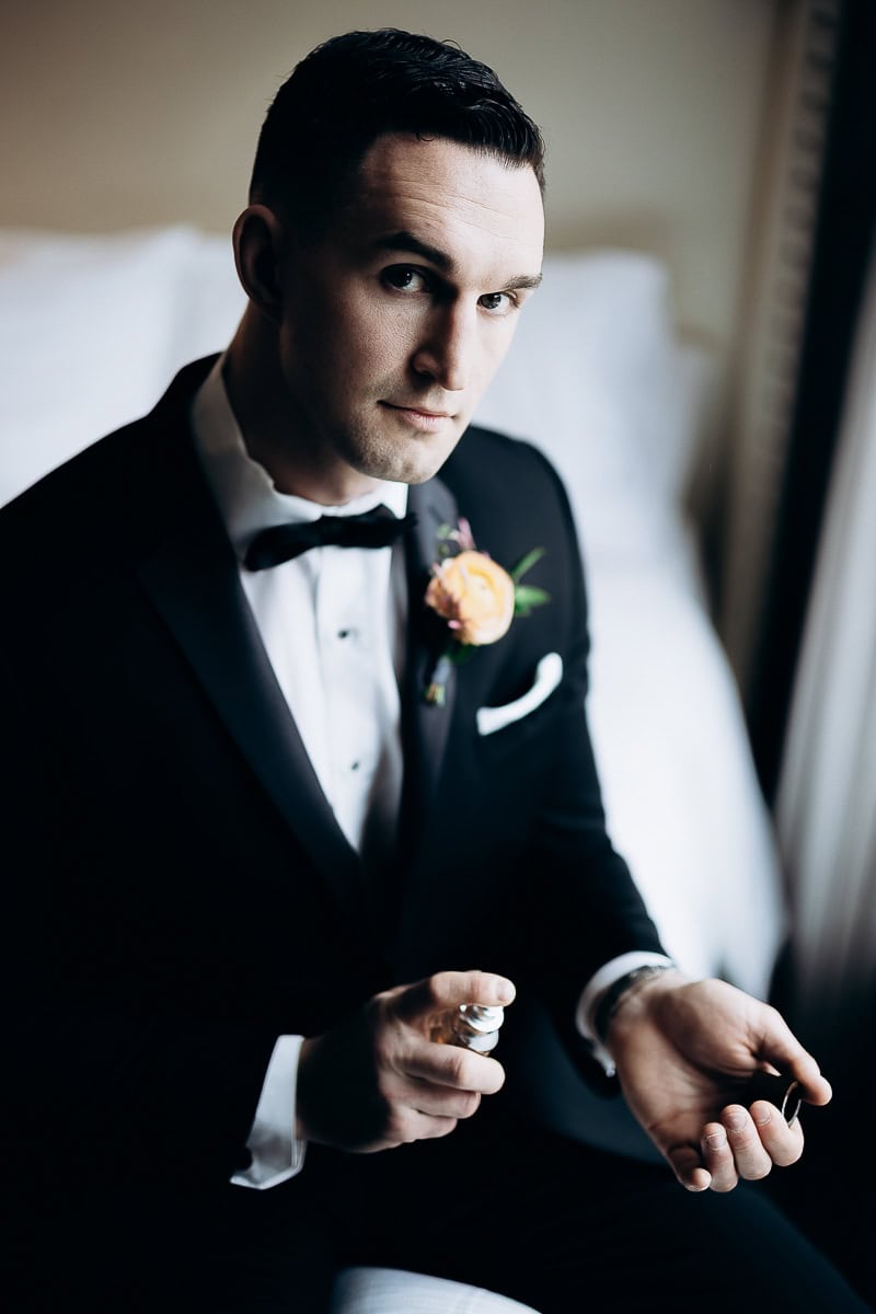 Man in black tuxedo with bow tie applying cologne on wrist, seated indoors near window light at The Stone House, Warren, NJ.
