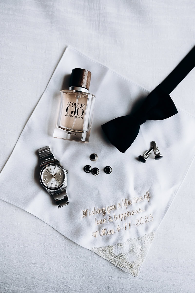 Groom's accessories including Acqua di Giò Absolu cologne, stainless steel watch, black bow tie, cufflinks, on embroidered handkerchief with wedding date.
