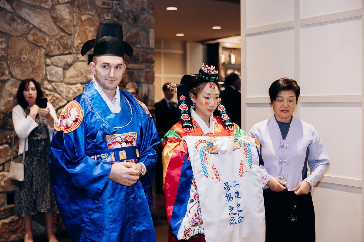 Bride and groom in traditional Korean wedding hanbok standing indoors at The Stone House, Warren, NJ, with a woman assisting the bride.