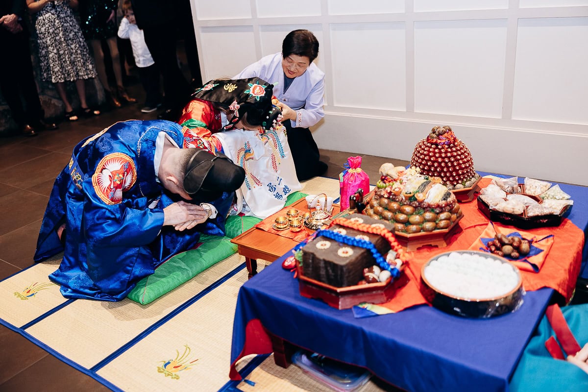 Couple in traditional Korean hanbok bowing deeply during paebaek ceremony next to a table with ceremonial foods and gifts at The Stone House, Warren, NJ.