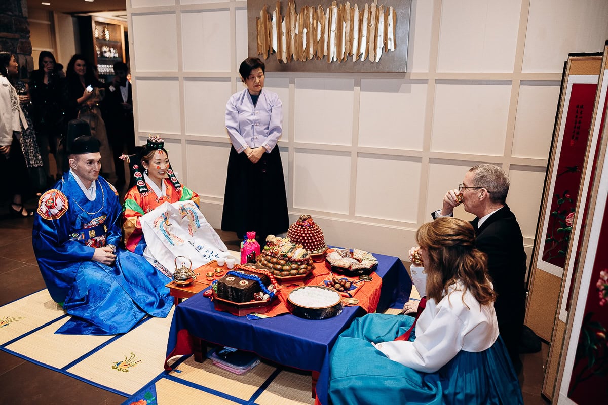 Couple in traditional Korean hanbok seated on mats during paebaek ceremony with ceremonial food and guests at The Stone House, Warren, NJ.