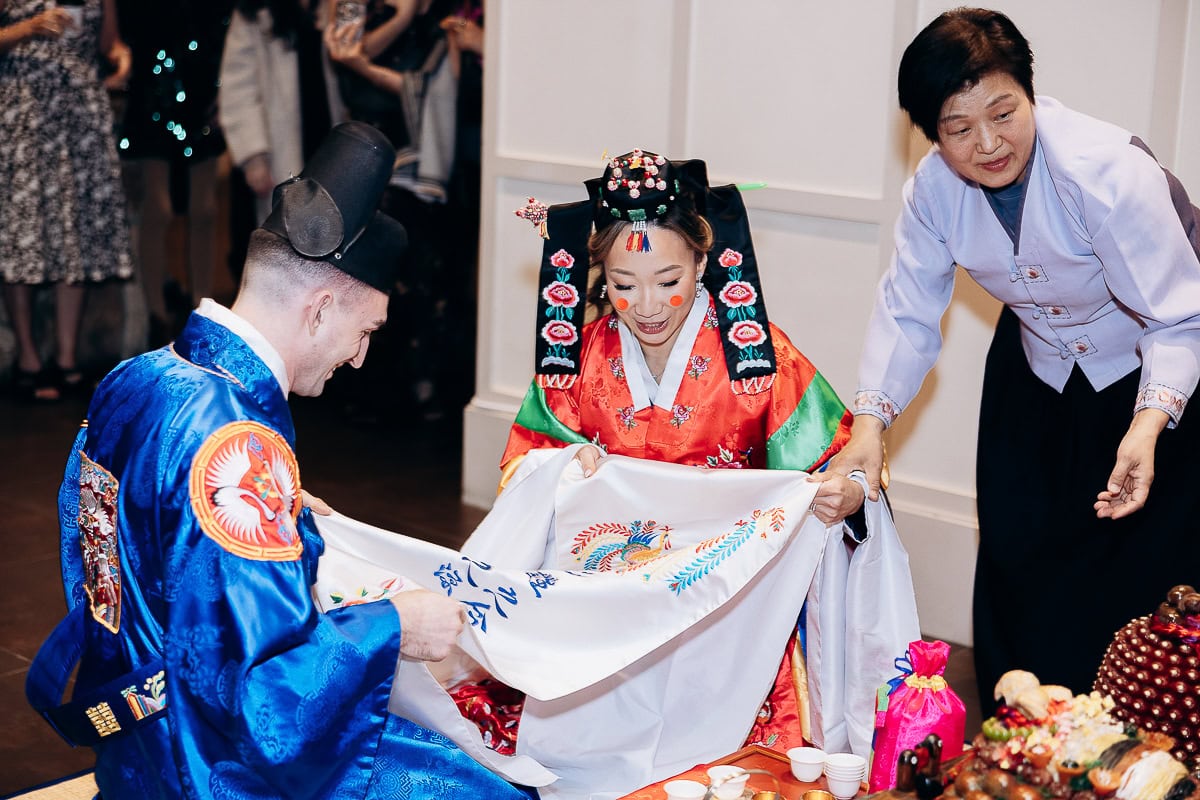 Couple dressed in traditional Korean hanbok performing paebaek ceremony with an elder assisting, surrounded by ceremonial items at The Stone House, Warren, NJ.