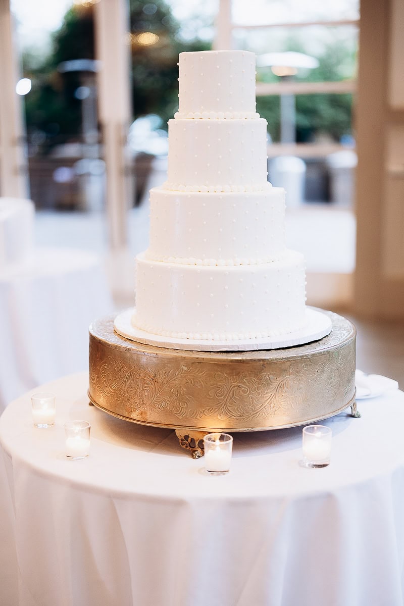 Four-tier white wedding cake with small pearl decorations on an ornate gold cake stand, placed on a white tablecloth with lit candles around it at The Stone House, Warren, NJ.
