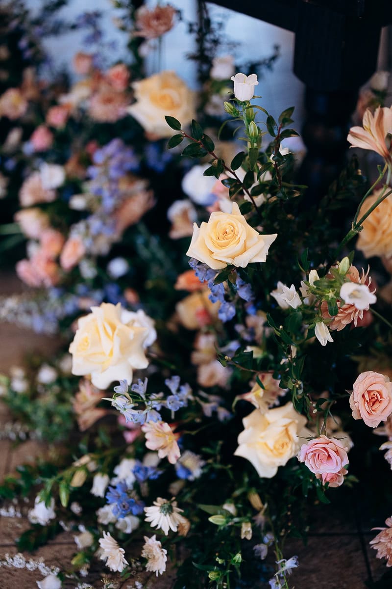 Close-up of a floral arrangement with cream roses, pink flowers, blue delphiniums, and green foliage on a stone floor at The Stone House, Warren, NJ.
