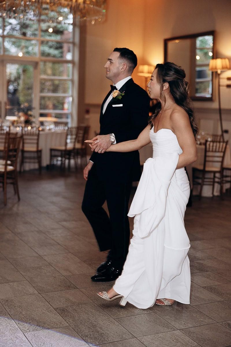 Bride in strapless white gown and groom in black tuxedo walking hand in hand through a softly lit reception room at The Stone House, Warren, NJ.