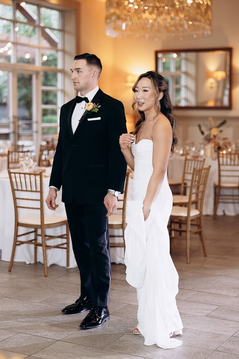 Bride in strapless white gown and groom in black tuxedo standing in a warmly lit wedding reception room with wooden chairs and tables at The Stone House, Warren, NJ.
