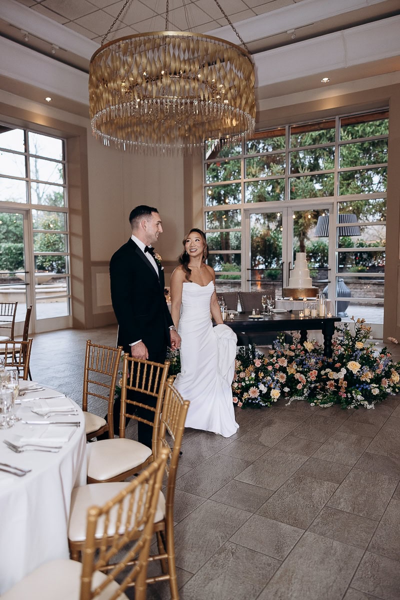 Bride in strapless white gown and groom in black tuxedo holding hands near floral sweetheart table with tiered cake, large chandelier overhead, and floor-to-ceiling windows at The Stone House, Warren, NJ.