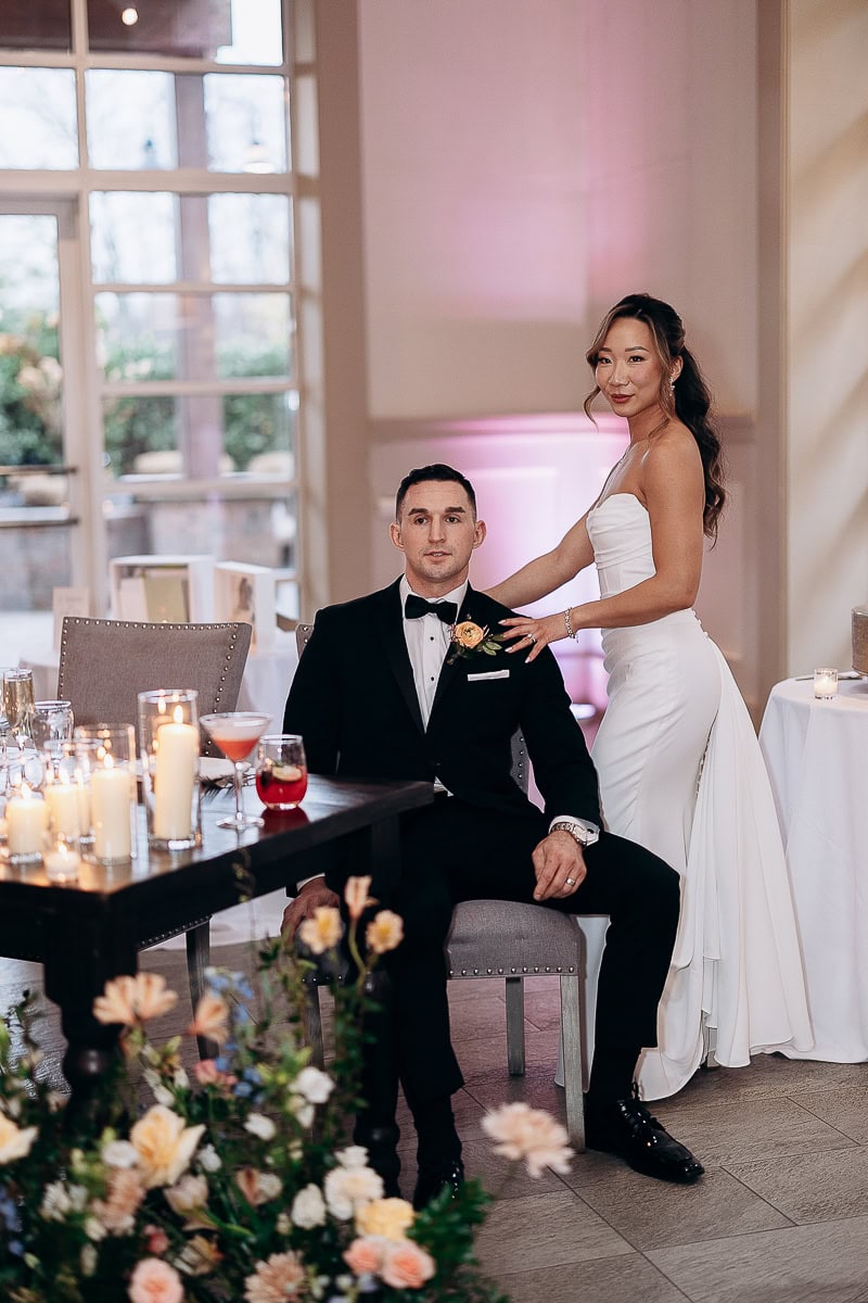 Bride in strapless white gown standing beside groom in black tuxedo seated at a candlelit table with floral arrangements at The Stone House, Warren NJ.
