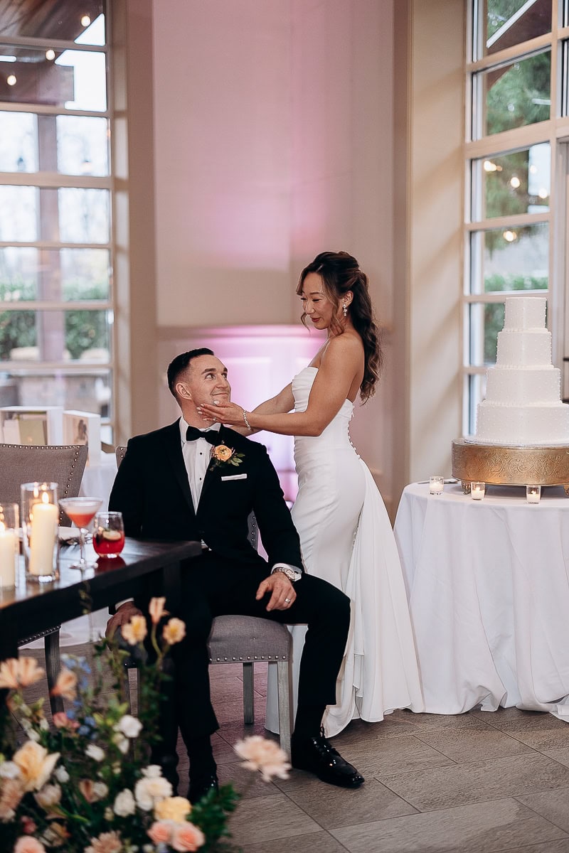 Bride in strapless white gown gently holding groom's face as he sits beside a white tiered wedding cake and lit candles indoors at The Stone House, Warren, NJ.