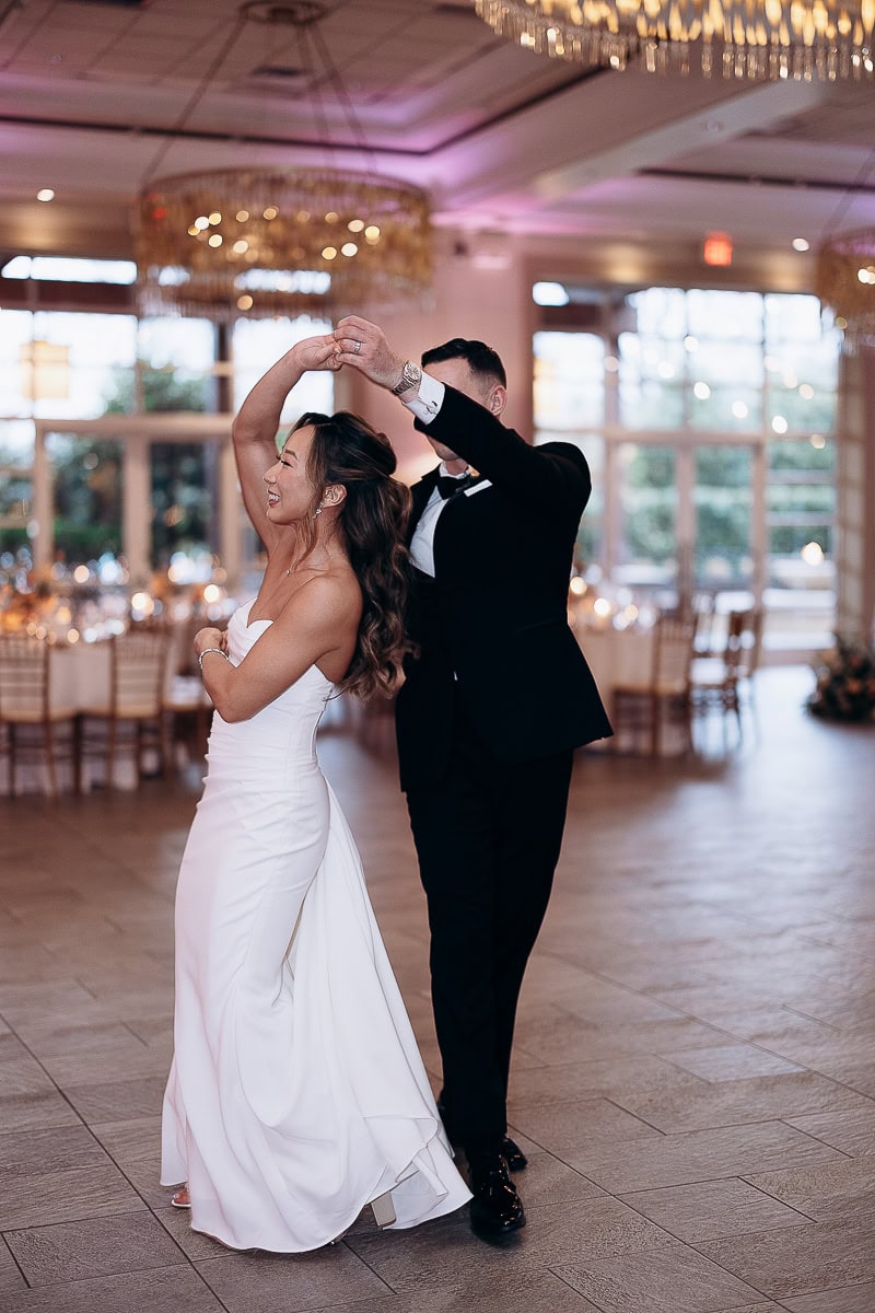 Bride in strapless white gown and groom in black tuxedo share a dance in a softly lit reception hall with chandeliers and round tables.