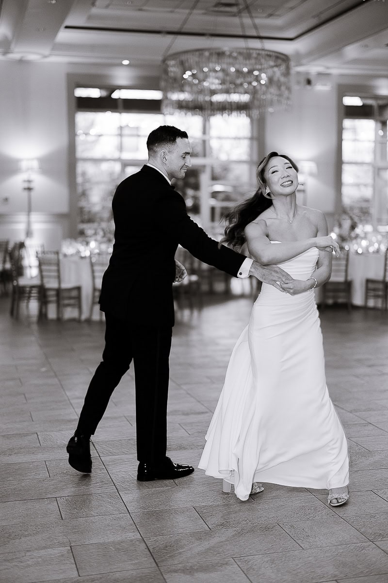 Black and white image of a couple dancing indoors, the groom in a classic black tuxedo and the bride in a strapless white gown, with a crystal chandelier and softly lit reception tables in the background at The Stone House, Warren, NJ.