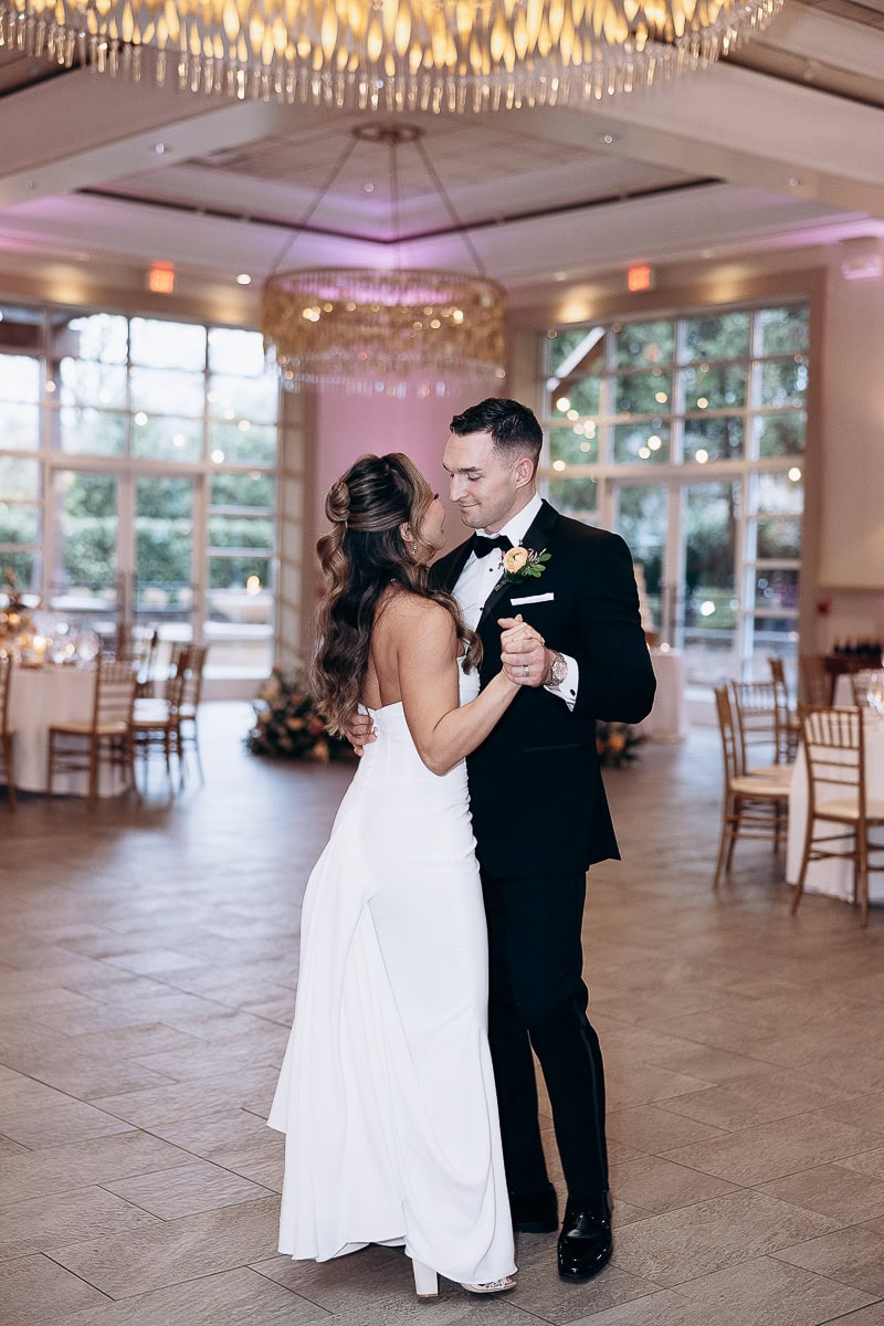 Bride and groom dancing closely in elegant wedding attire in a softly lit reception hall with large windows and chandeliers at The Stone House, Warren, NJ.
