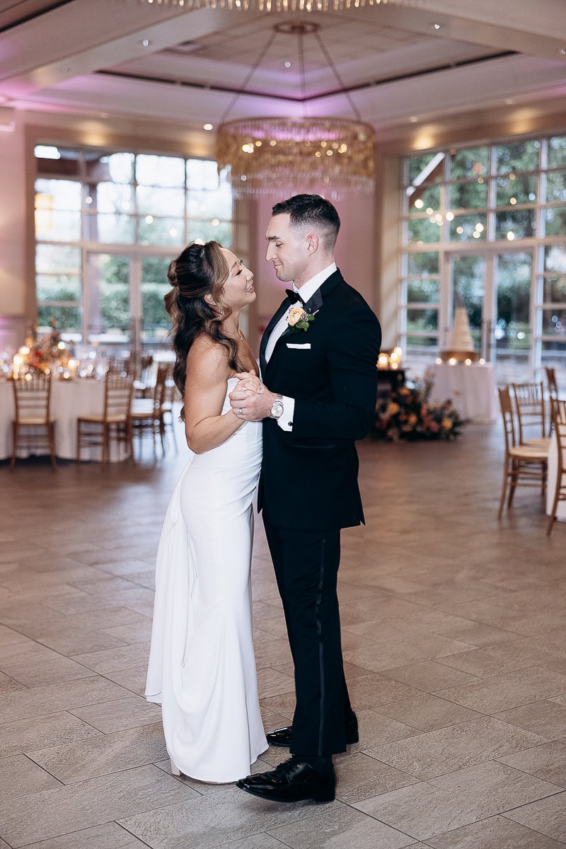 Bride and groom dancing closely in a softly lit reception hall with large windows and a crystal chandelier at The Stone House, Warren, NJ.