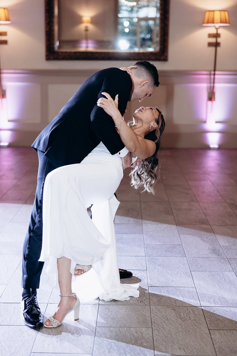 Groom in black suit dipping bride in white dress during wedding dance at The Stone House, Warren, NJ, with warm ambient lighting and tiled floor.