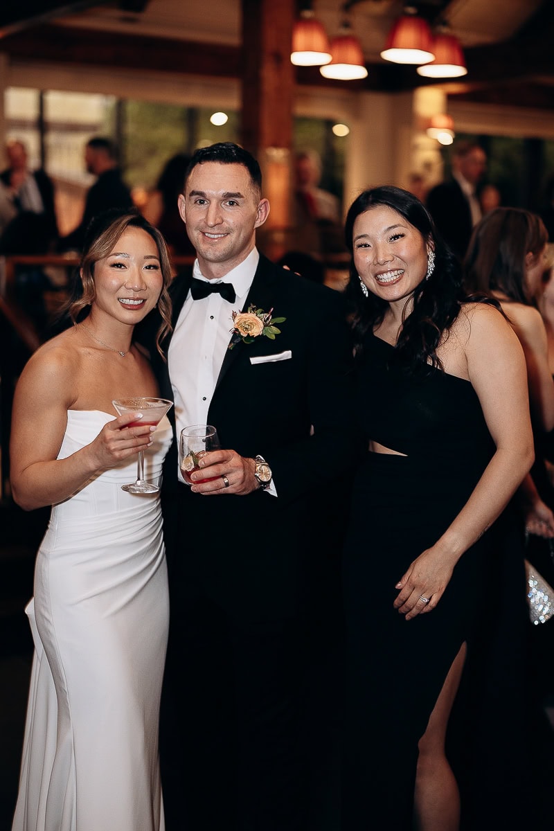 Three elegantly dressed guests at a wedding reception at The Stone House, Warren NJ, holding cocktails and smiling in warm ambient lighting.
