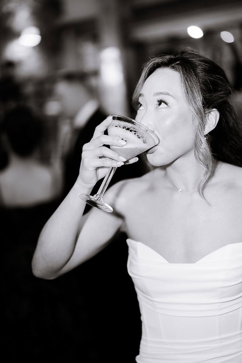 Bride in strapless white dress drinking from a cocktail glass, softly lit with blurred guests in background at wedding reception.