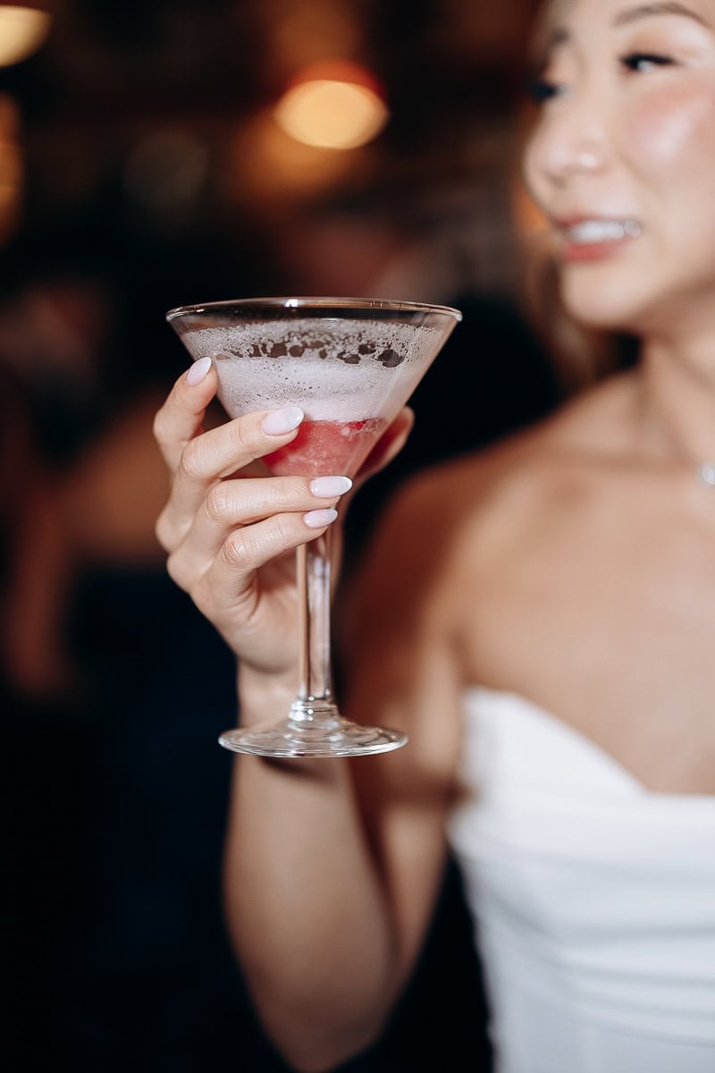 Close-up of a woman in a white strapless dress holding a frosted cocktail glass with a red drink inside, softly blurred background.