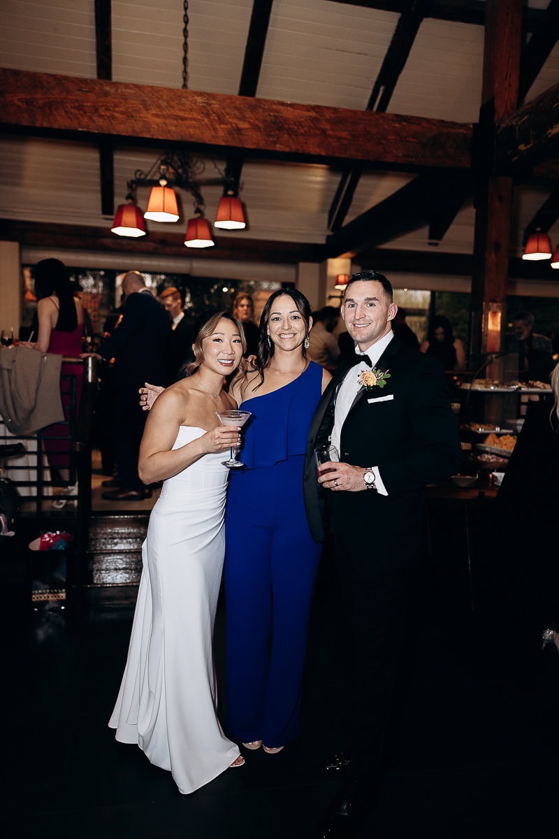 Three elegantly dressed guests at a wedding reception inside The Stone House, Warren, NJ, with wooden beams and warm pendant lighting overhead.