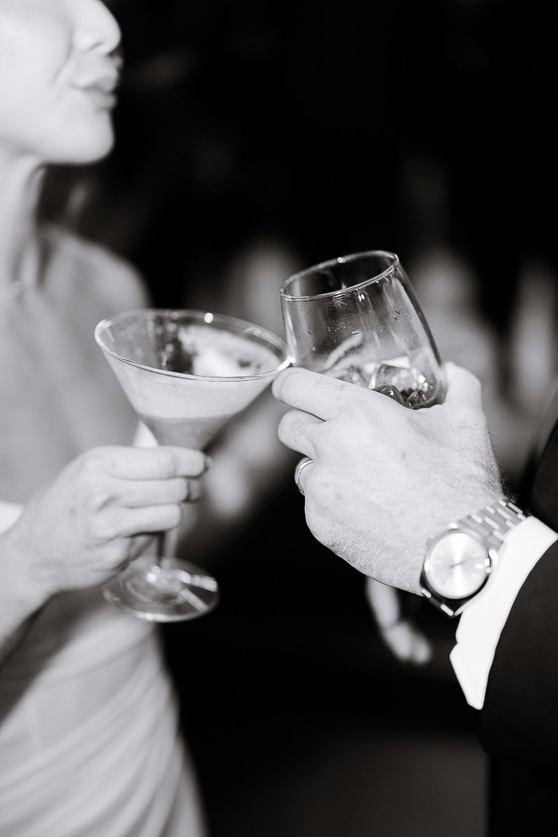 Close-up of two hands holding cocktail glasses mid-toast, one with a martini glass and the other with a tumbler, set against a blurred background.