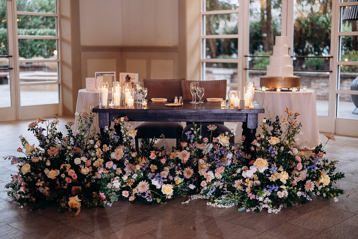 Sweetheart table with dark wood, surrounded by pastel floral arrangements and lit candles, with a tiered wedding cake in the background at The Stone House, Warren, NJ.