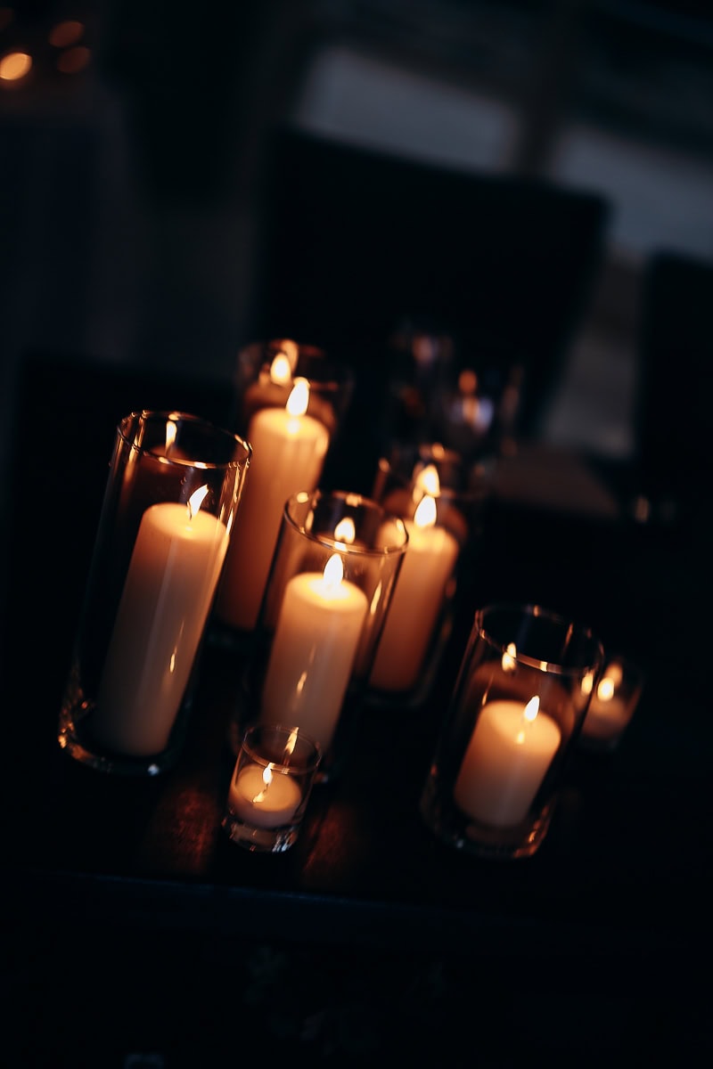 Cluster of lit white pillar candles in clear glass holders on a dark surface, creating a soft, warm ambiance at The Stone House, Warren, NJ.