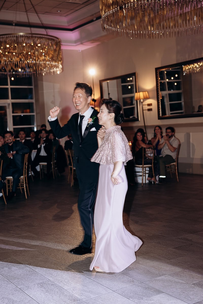 An elegantly dressed couple dancing in a softly lit reception hall with crystal chandeliers and seated guests applauding in the background at The Stone House, Warren, NJ.